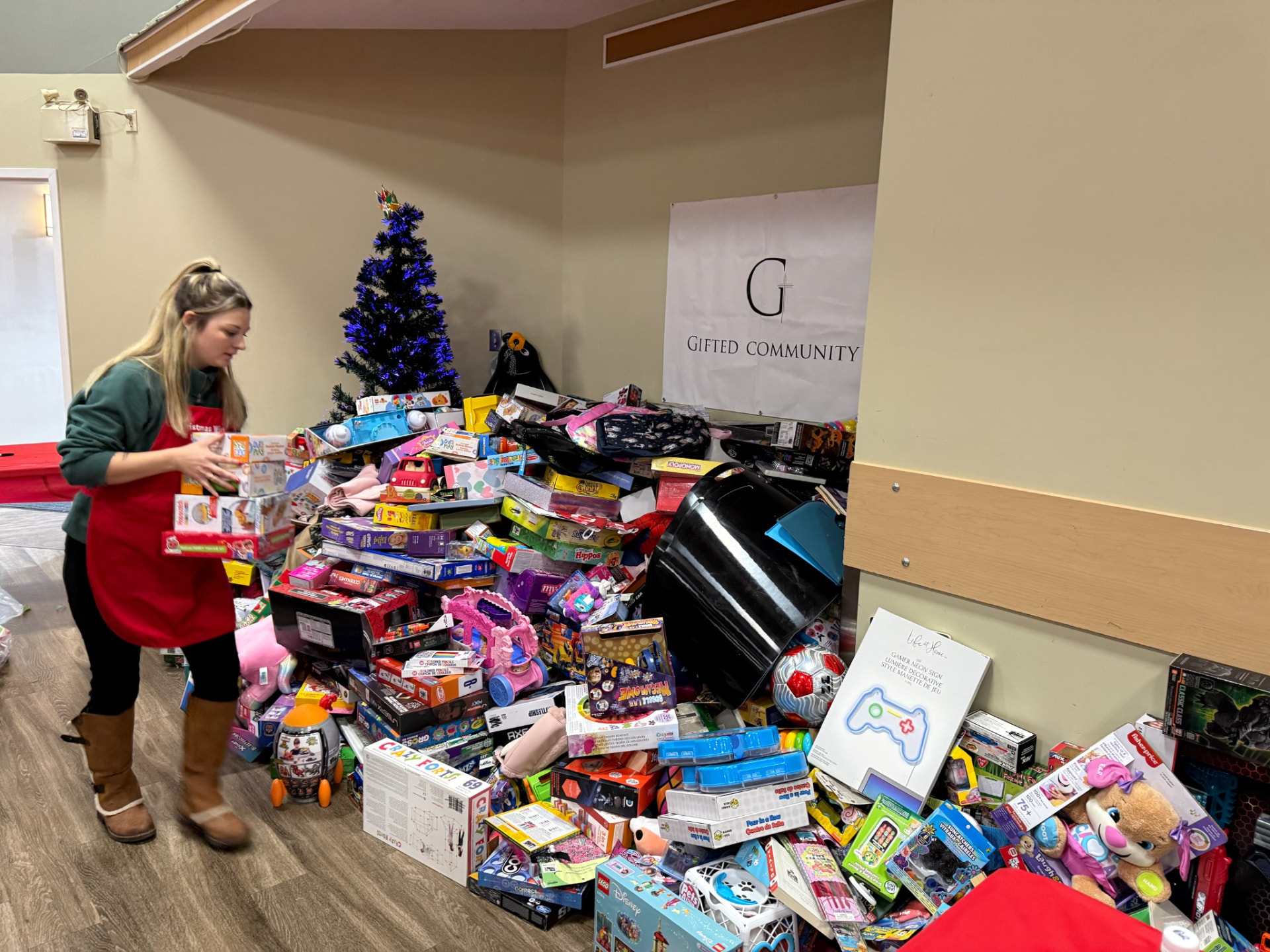 A volunteer in a red apron sorting donated gifts beside a Christmas tree